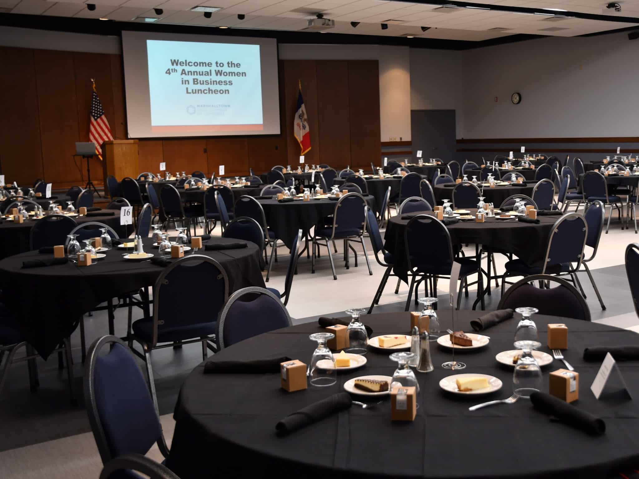 Conference center with tables set for an event facing a dais with a projection screen for presentations.